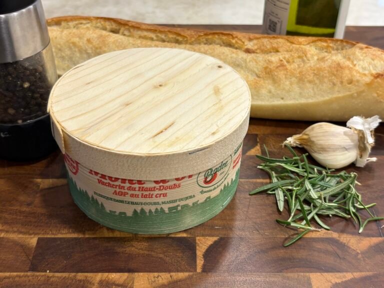 A wooden box of Mont d'Or cheese next to rosemary, garlic, pepper, and wine.